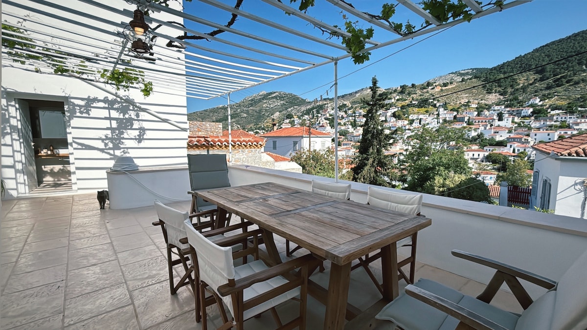 An outdoor terrace is featured, showcasing a wooden dining table surrounded by several chairs. Sunlight filters through a pergola, highlighting the surrounding views of hillside homes and greenery. The area is spacious, providing ample room for relaxation or dining.
