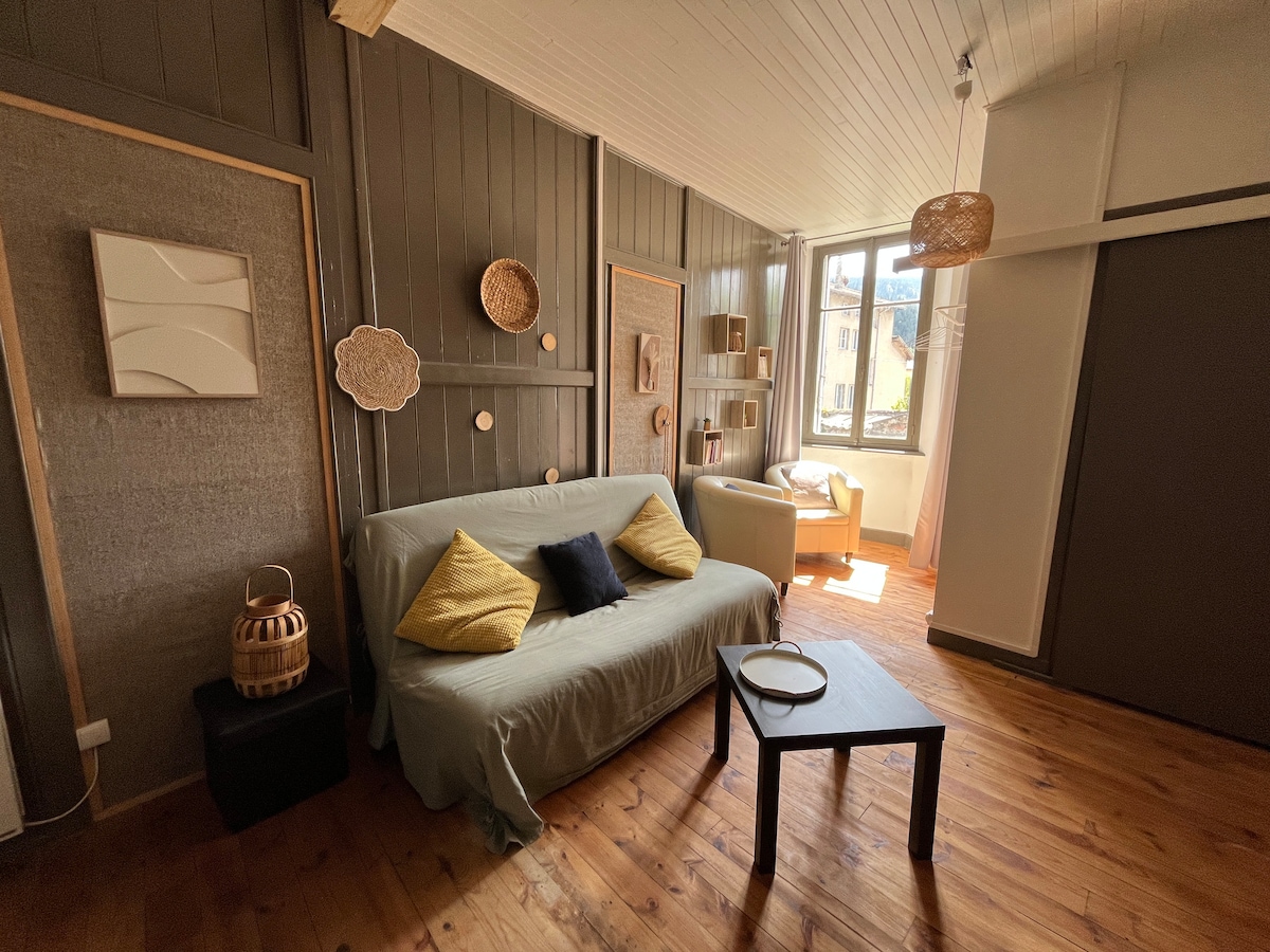 A living area features a light-colored sofa adorned with decorative cushions, complemented by a small wooden coffee table. Natural light filters through a window, highlighting the wooden flooring and textured wall elements, while woven wall decorations add visual interest to the space.