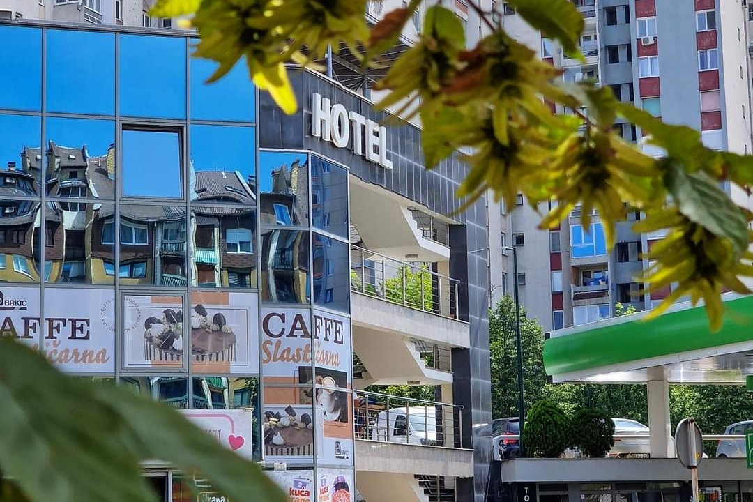 The hotel exterior is showcased, featuring a modern glass facade reflecting surrounding buildings and greenery. A sign displaying the word 'HOTEL' is positioned prominently above the entrance. Lush green leaves frame the image in the foreground, adding a natural touch to the urban setting.