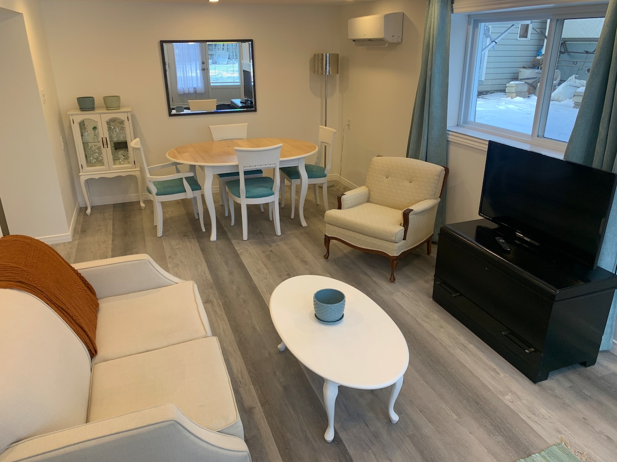 A bright living space is shown, featuring a comfortable seating arrangement with a light-colored sofa and armchair. A round dining table with blue chairs is positioned nearby. A television stands on a black console, and natural light enters through a window with light curtains.