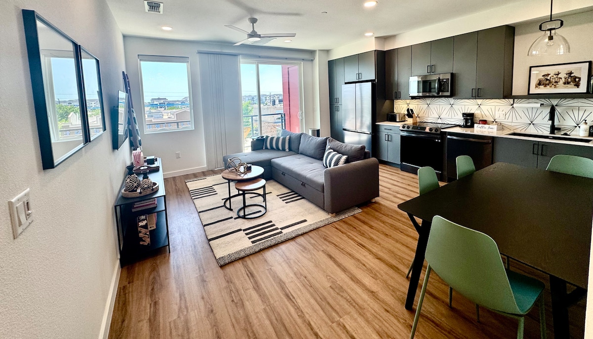 A spacious living area integrates a comfortable couch with a coffee table, complemented by a decorative rug. Nearby, a modern dining table with green chairs is visible. Large windows allow natural light to fill the space, offering views of the surroundings. The kitchen features dark cabinets and sleek appliances.