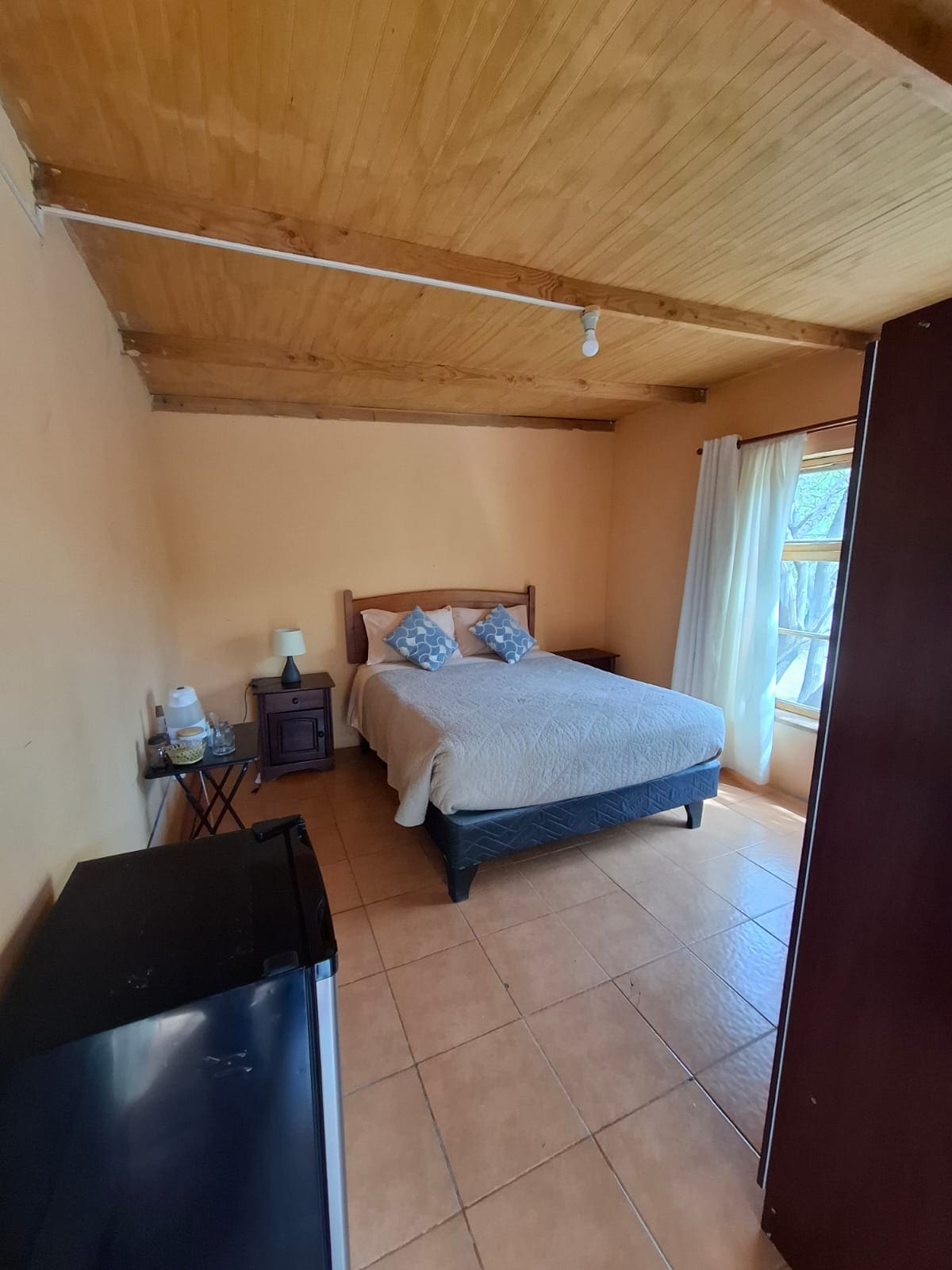 A spacious sleeping area is presented with a neatly made bed featuring a light-colored quilt. Two decorative cushions adorn the headboard, while a bedside table with a lamp is visible. A window with sheer curtains allows natural light to enter, highlighting the warm-toned walls and tiled floor.