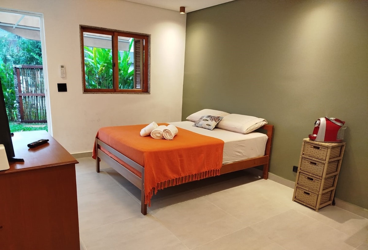 A cozy sleeping area features a wooden bed with an orange blanket laid across it. A small table houses a television. Natural light enters through a window with a view of lush greenery outside. A wicker basket stands beside the bed, enhancing the room's functionality.