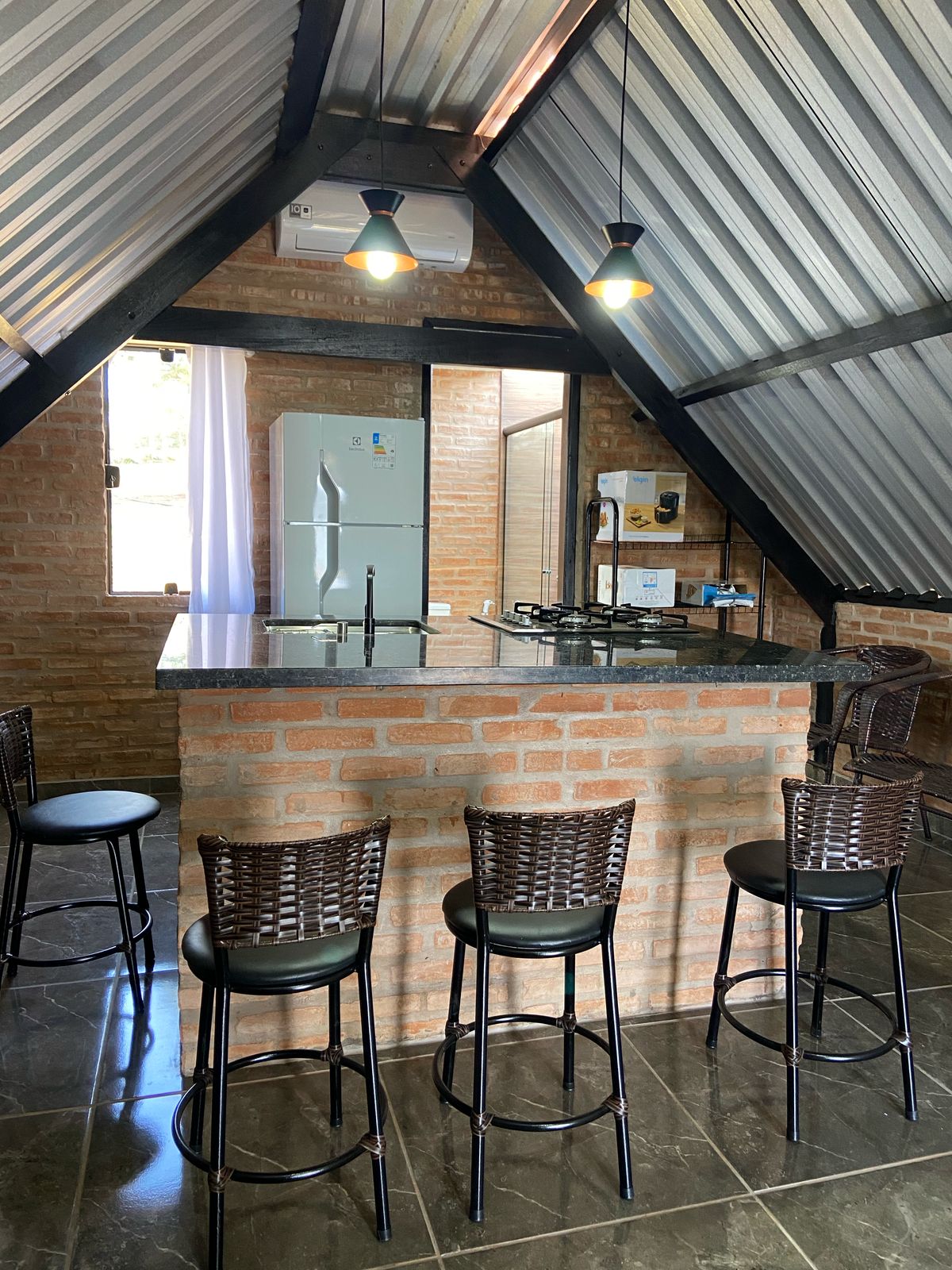 The kitchen area features a central island bar with four high stools arranged around it. A sleek countertop complements the modern appliances, including a refrigerator and stove. Large windows allow natural light to fill the space, showcasing the interior's rustic brick walls.