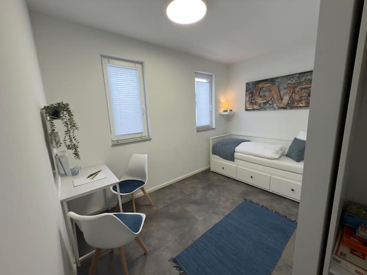 Chumba rahisi cha kulala kilicho na dawati, kitanda na mkusanyiko wa michezo ya ubao kwenye kabati.