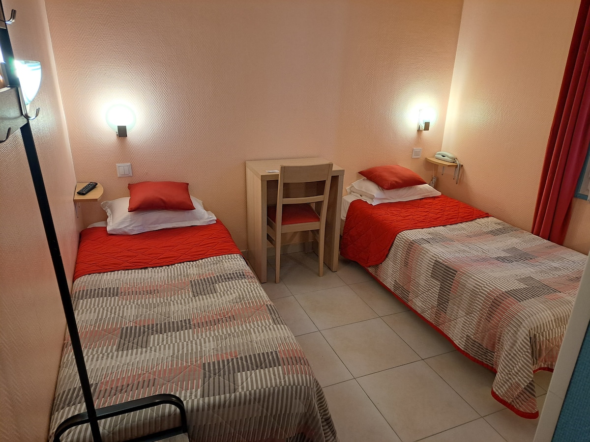 The room features two twin beds with red bedspreads and patterned blankets. A simple desk and chair are positioned between the beds. Soft lighting is provided by wall-mounted fixtures, while tiled flooring offers a clean and functional surface.
