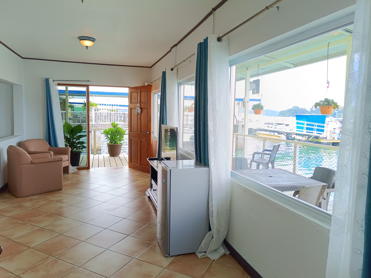 A bright and airy living space features large windows allowing natural light to illuminate the tiled floor. A cozy seating area with a chair and potted plant complements the space. A small fridge and television provide convenience, while views of the outside are visible.
