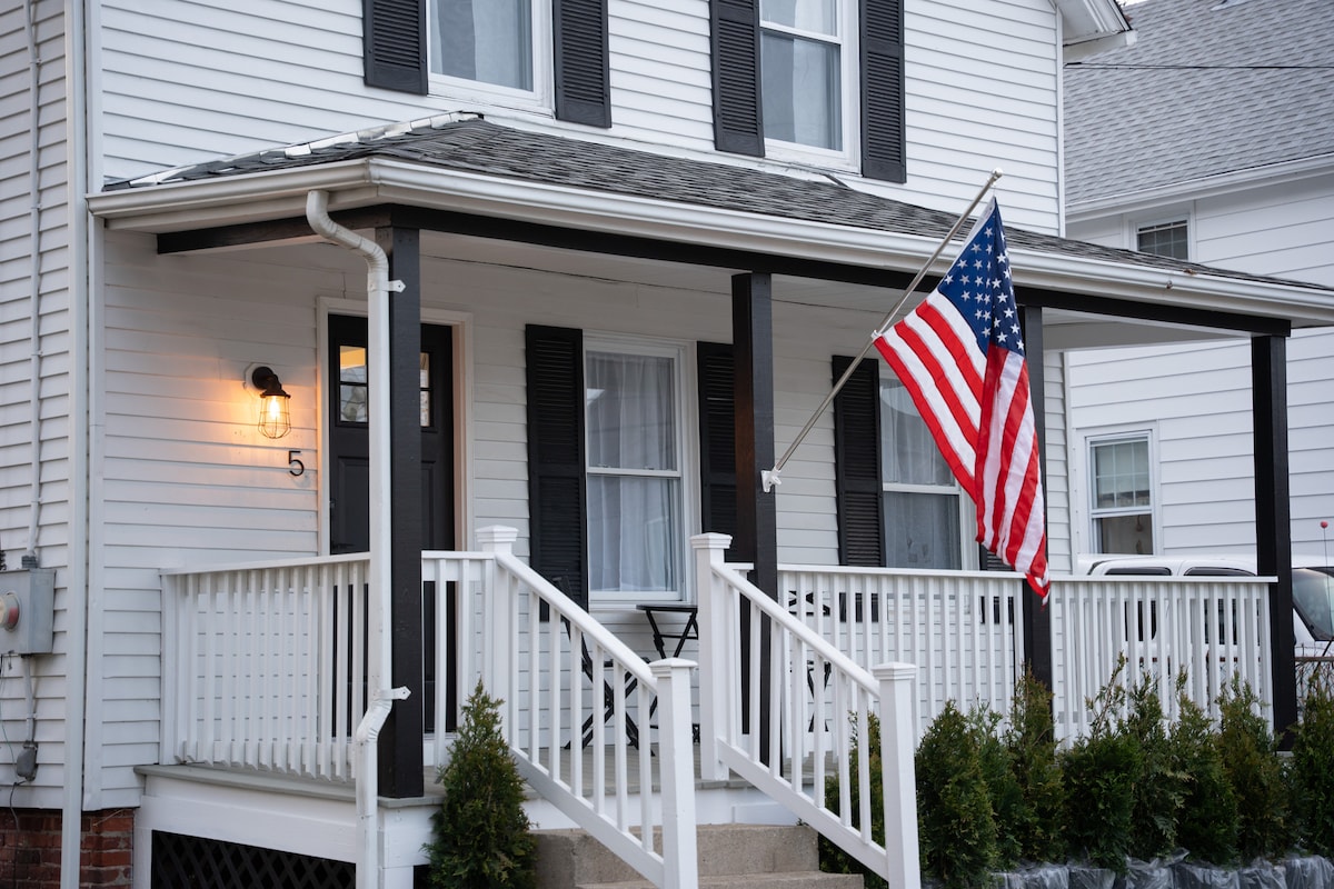 The exterior of a renovated New England home features a welcoming front porch with a white railing, complemented by an American flag. Large windows are flanked by black shutters, and neatly trimmed greenery lines the base, providing a cozy entrance.