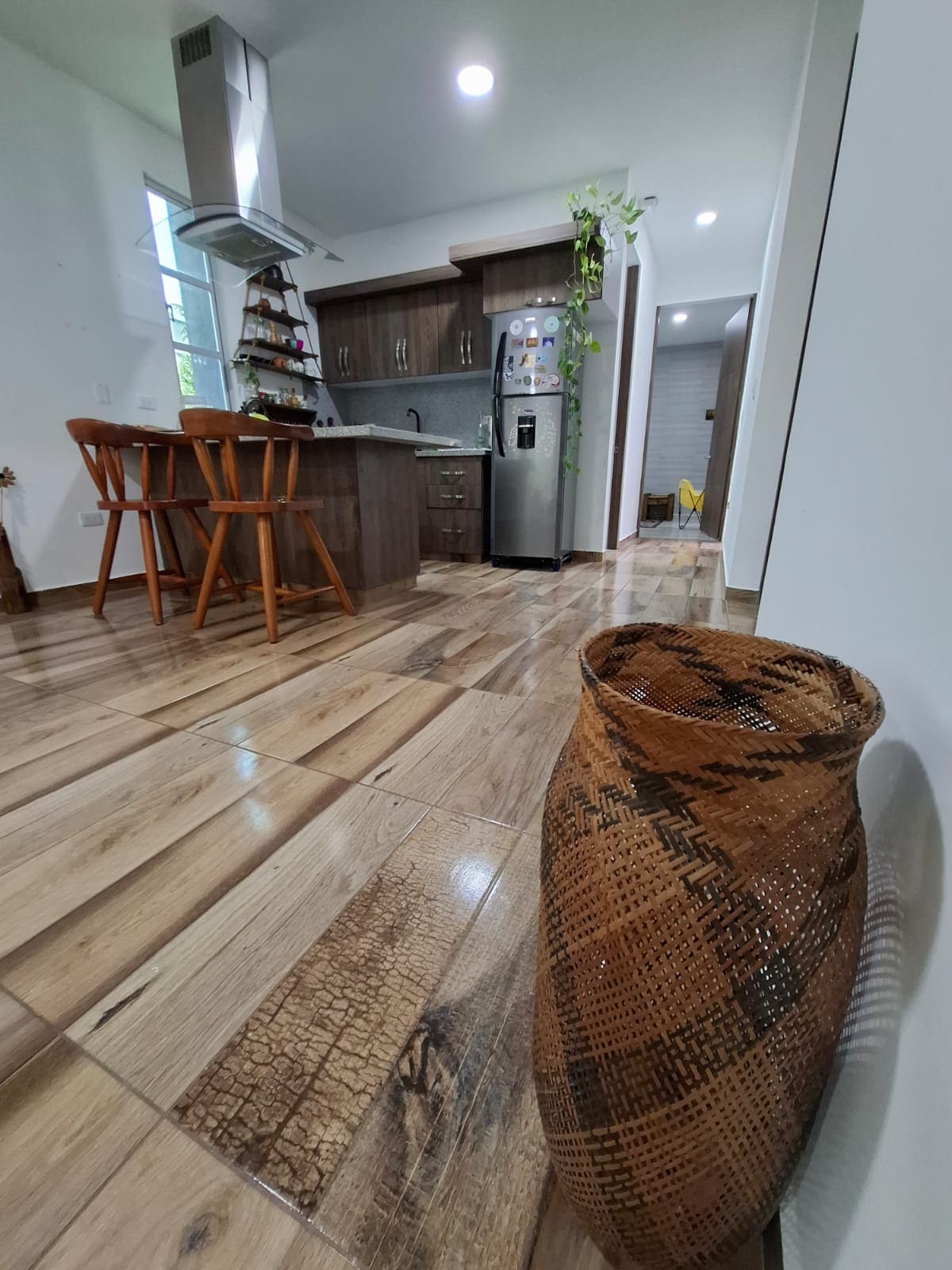 A modern kitchen area is presented, featuring wood cabinetry and stainless steel appliances. Two wooden bar stools sit at a counter. The flooring showcases a blend of light and dark wood tones, while a woven vase adds an element of texture near the foreground.