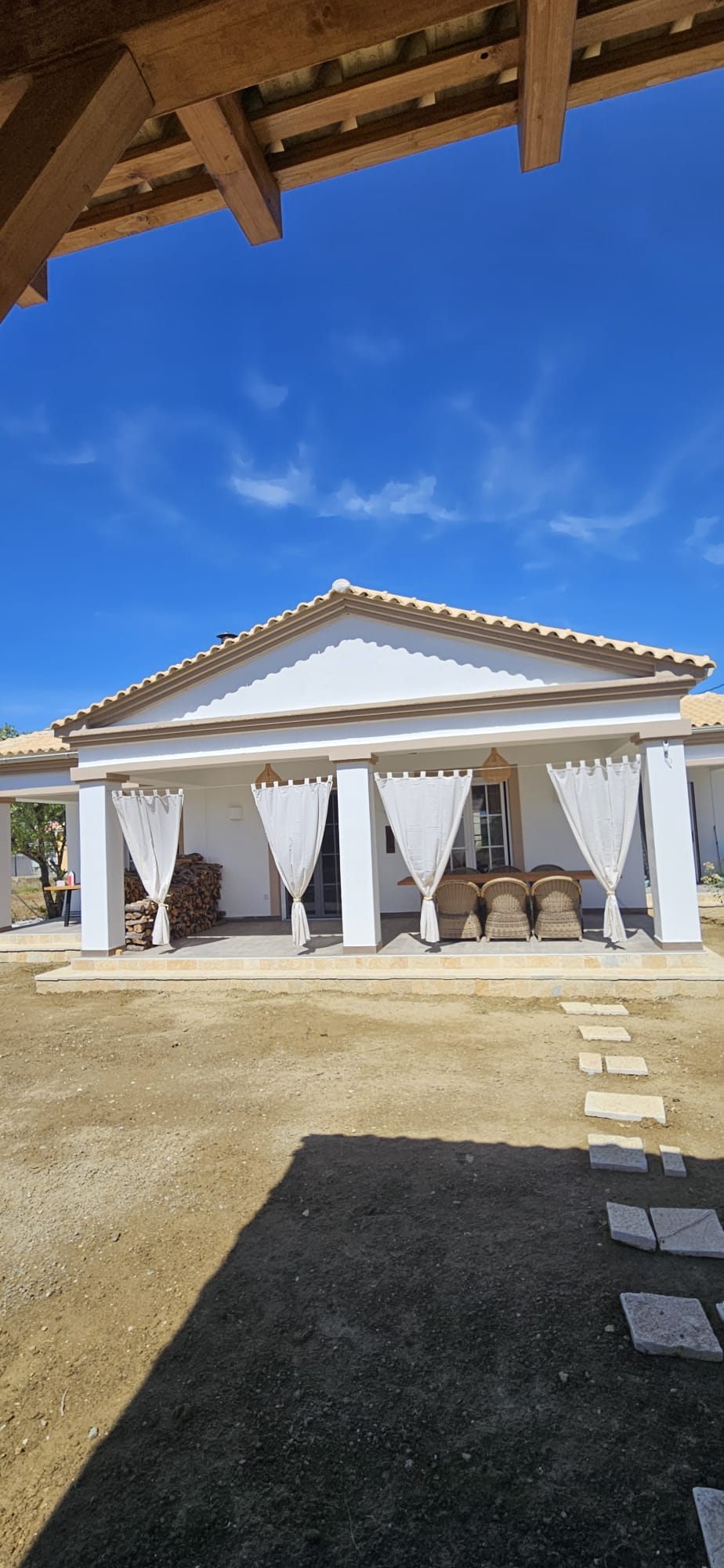 The exterior of the villa displays a white facade with a traditional roof design, complemented by two decorative pillars. Sheer curtains frame the entrances, and a pathway of stepping stones leads to the porch area. The sky above is clear and bright, enhancing the overall setting.