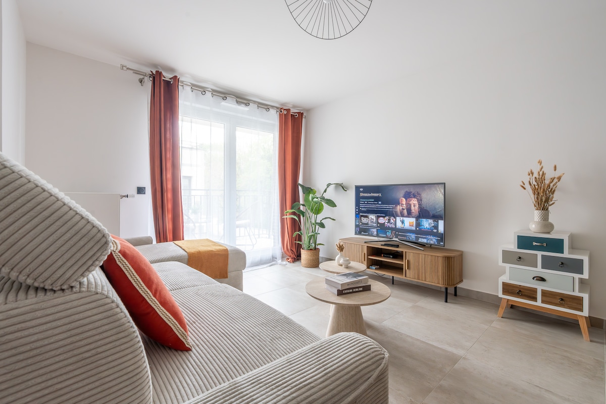 A modern living area features a comfortable sofa with accent pillows, a round coffee table, and a television on a stylish media unit. Large windows invite natural light, while orange curtains add a touch of warmth. A potted plant enhances the inviting atmosphere.