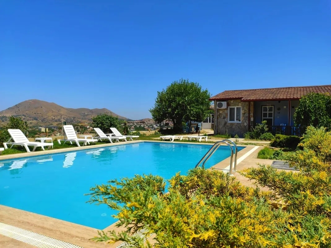 A clear blue pool is framed by sun loungers and lush greenery, offering a serene outdoor space. A modest building with a shaded porch is visible nearby, set against a backdrop of rolling hills under a bright blue sky.