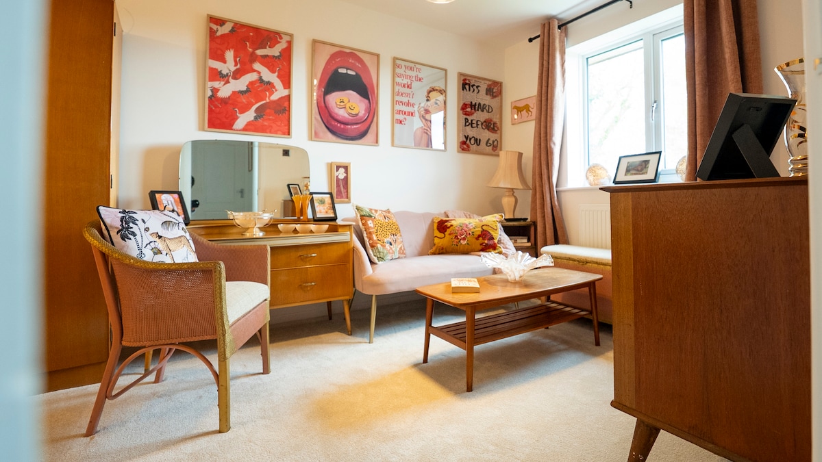 A cozy living area features a soft carpet underfoot and a selection of mid-century modern furniture. A pink sofa with decorative cushions is complemented by two chairs. Colorful artwork adorns the walls, while a wooden sideboard adds character. Natural light fills the space through a large window.