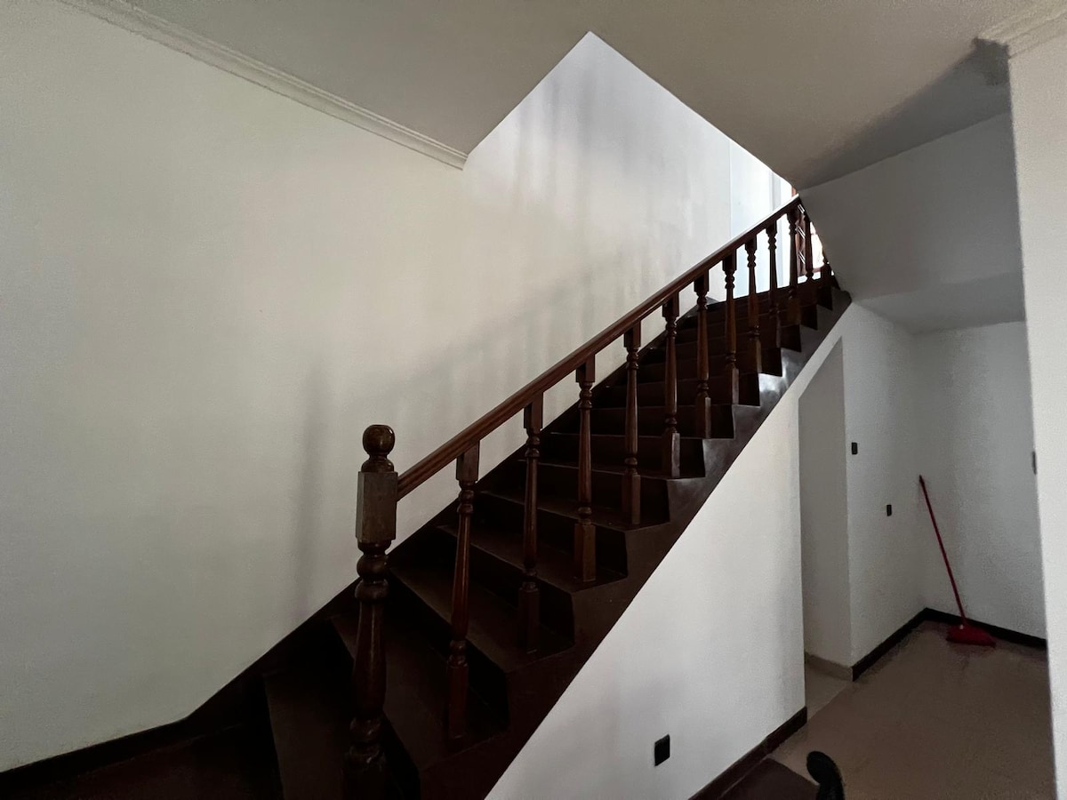 A wooden staircase is seen ascending along a light-colored wall. The railings are designed with rounded balusters, and the staircase has a dark finish, contrasting with the neutral tones of the surrounding area. Natural light is cast on the steps.