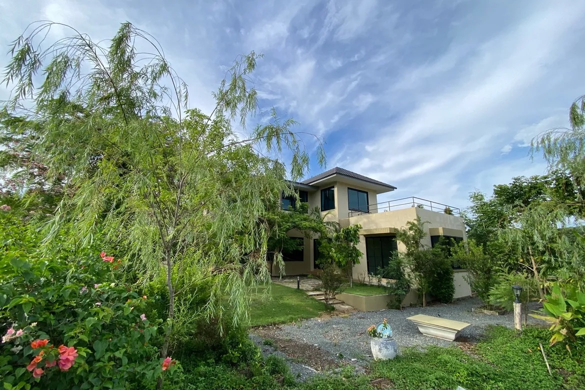 A two-story modern house is surrounded by lush greenery, featuring a variety of mature trees and flower bushes. The soft landscaping includes a gravel pathway leading to the entrance. The sky above is partly cloudy, enhancing the peaceful atmosphere of the outdoor space.