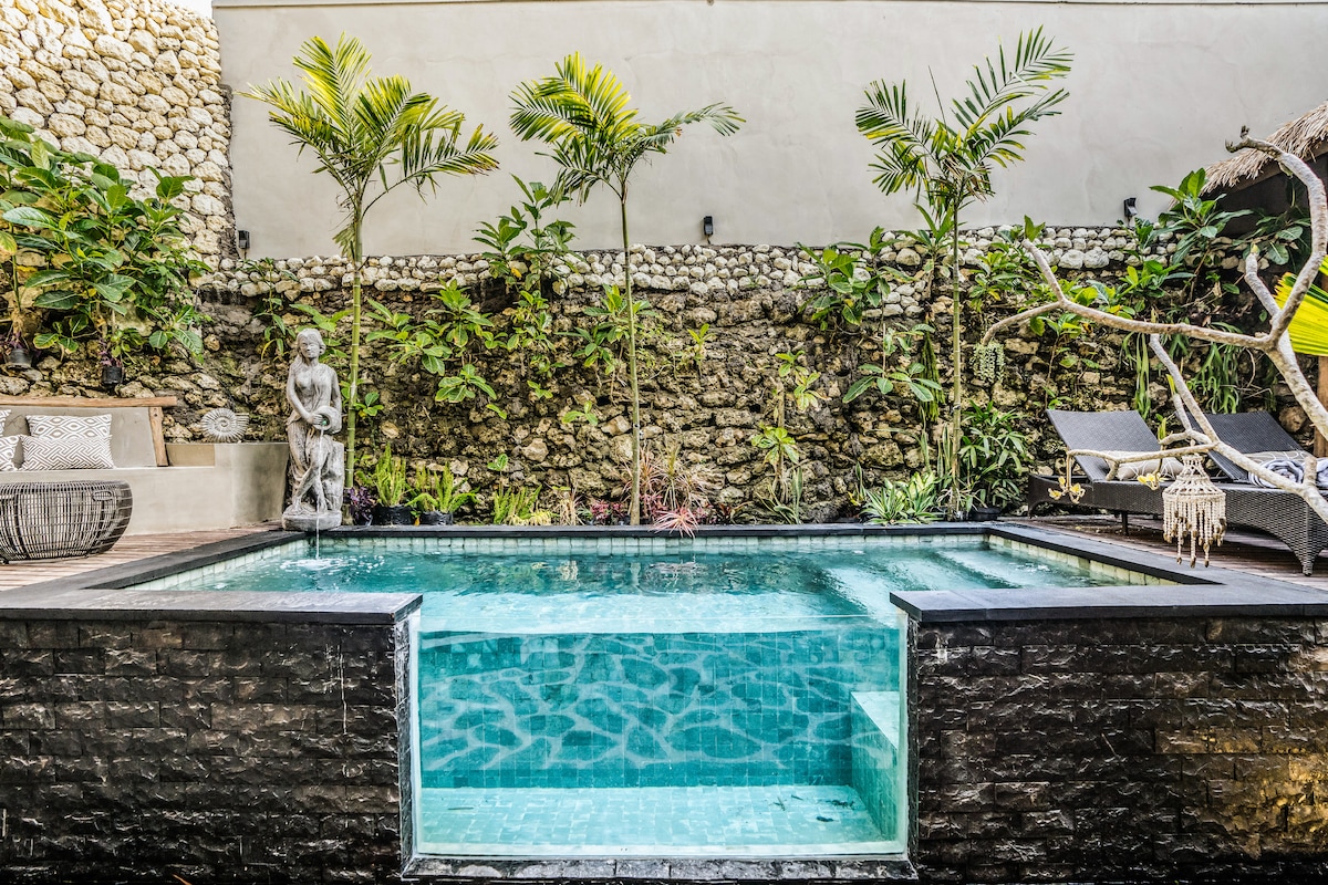 A tranquil pool is surrounded by a stone wall and lush tropical plants, including palm trees. A decorative statue is positioned near the pool's edge, adding a serene touch to the outdoor space.