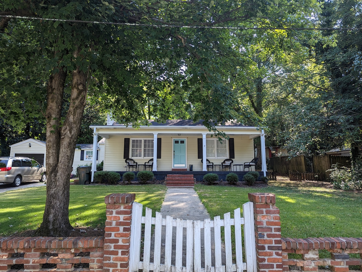 A charming single-story home is set behind a white picket fence, featuring a welcoming porch with black rocking chairs. The light yellow exterior is complemented by aqua-colored front door. Mature trees provide shade to the yard, which is well-kept and lined with manicured bushes.