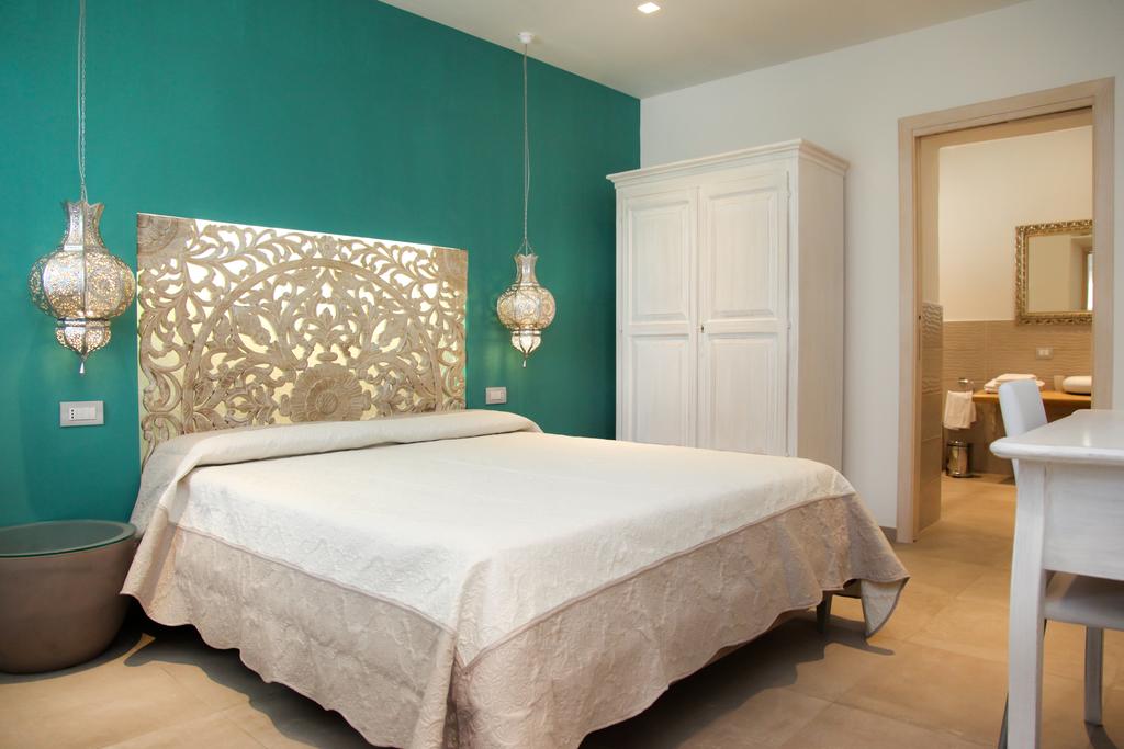 A spacious bedroom features a light-toned bed with a decorative headboard, complemented by intricately designed hanging lanterns. The walls are painted a soothing teal, and a white wardrobe is positioned nearby. An archway leads to an adjoining area, hinting at additional amenities.