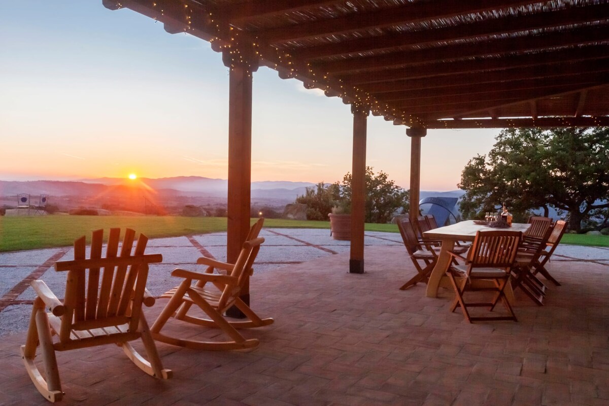 A covered outdoor patio area features wooden rocking chairs and a dining table, positioned to capture the sunset view over the distant hills. Soft lighting hangs above, enhancing the serene atmosphere as the sun sets beyond the horizon.