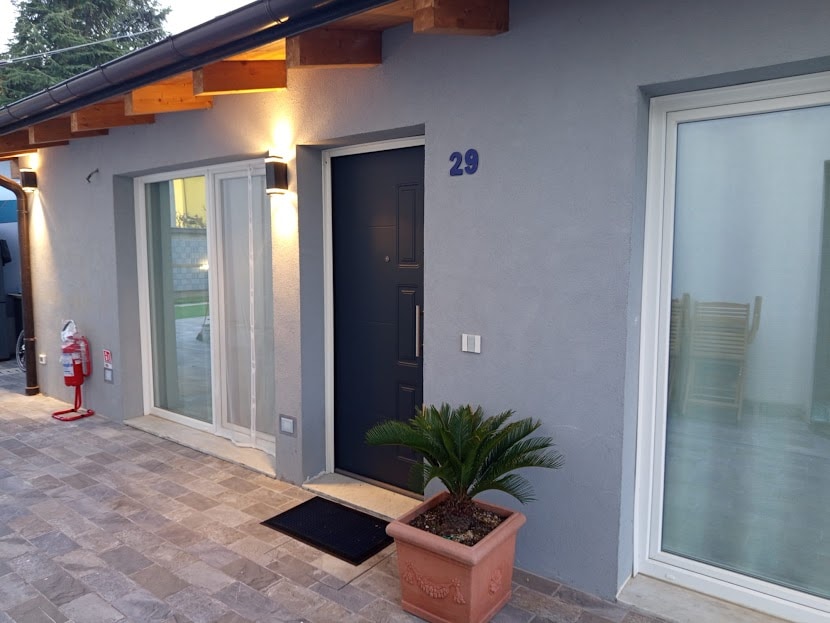 The entrance of the independent studio features a modern door, numbered 29, framed by large windows. A planter with greenery stands on the doorstep, alongside a welcoming mat. Soft lighting enhances the structure's clean lines, while the stone pathway adds to the inviting approach.