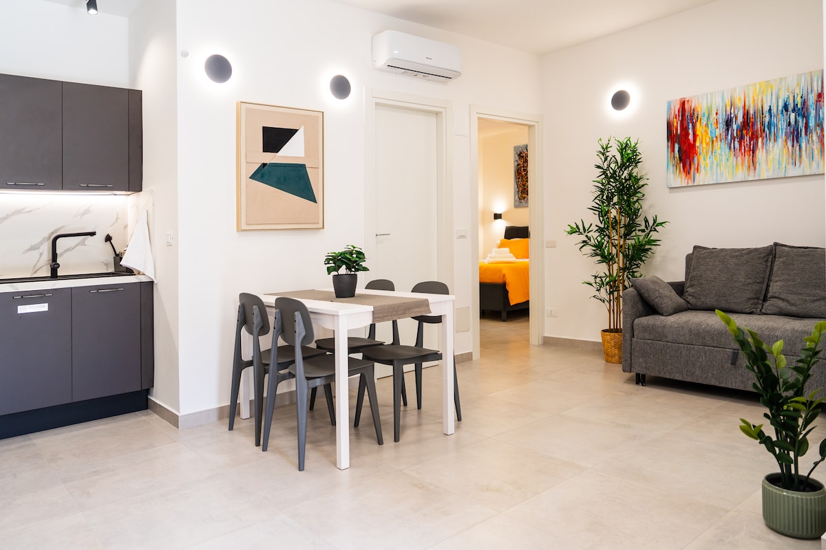 An open living area features a modern kitchen with dark cabinetry adjacent to a dining table, accompanied by four chairs. A grey sofa sits in the corner, with abstract wall art adding color. A doorway leads to a brightly lit bedroom beyond.