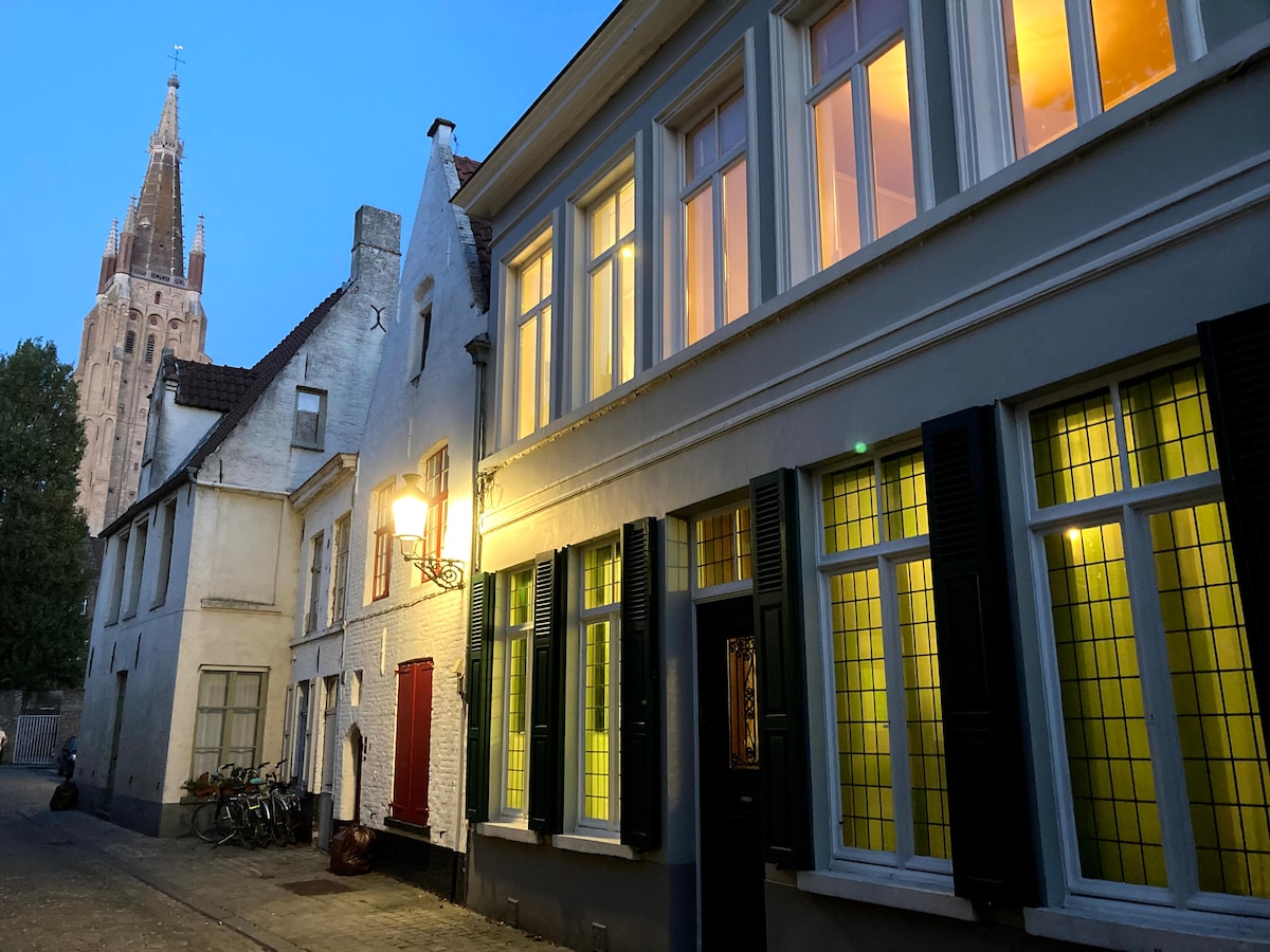 An evening view of a charming street in Brugge, featuring a historic building with large windows illuminated in warm tones. A traditional lamp post casts a soft glow near the entrance, while the silhouette of a church tower is visible in the background.