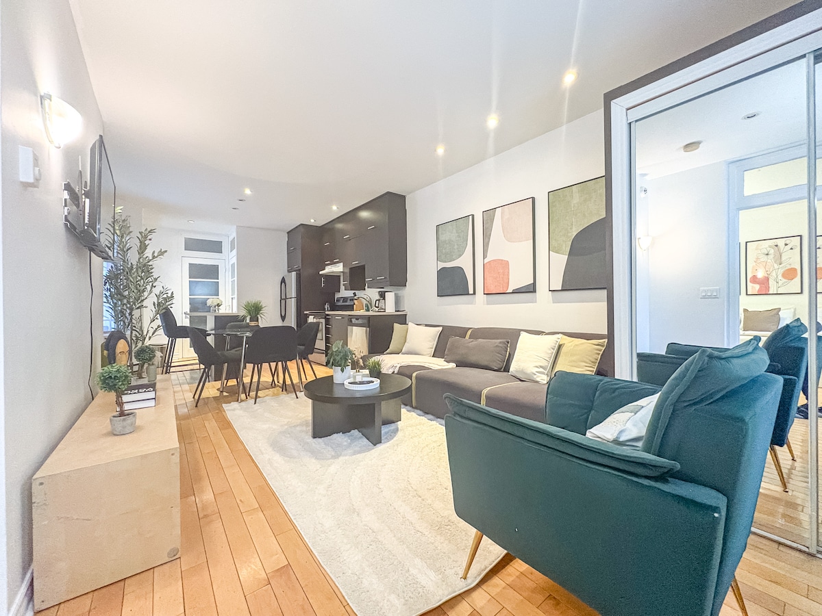 A modern living area is furnished with a spacious sectional sofa in a neutral tone and a stylish armchair. A round coffee table centers the space, surrounded by a dining set. Artwork decorates the walls, and natural light fills the room from large windows.
