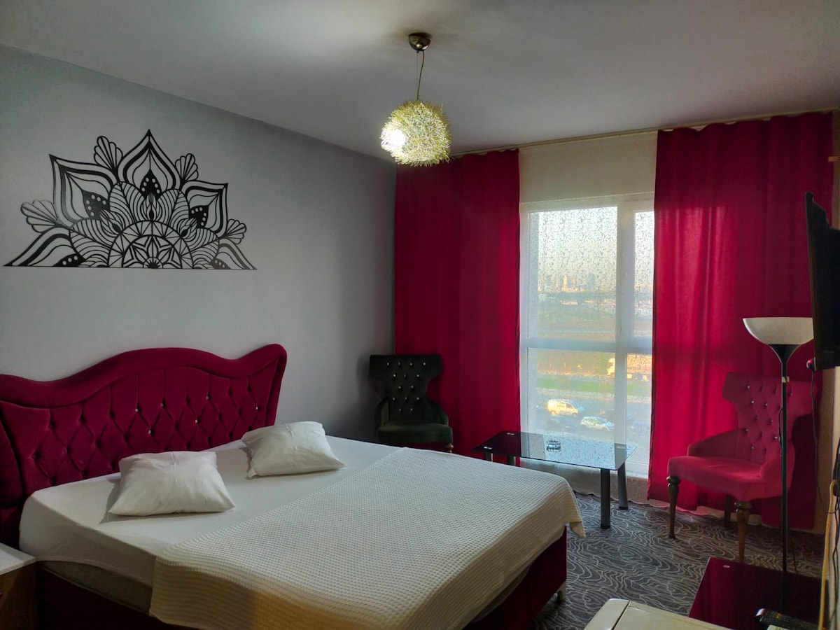 A spacious bedroom features a large bed with two pillows and an elegant headboard. Light is filtered through a large window adorned with sheer curtains, while rich red drapes frame the window. Two armchairs and a glass table complete the inviting setting.