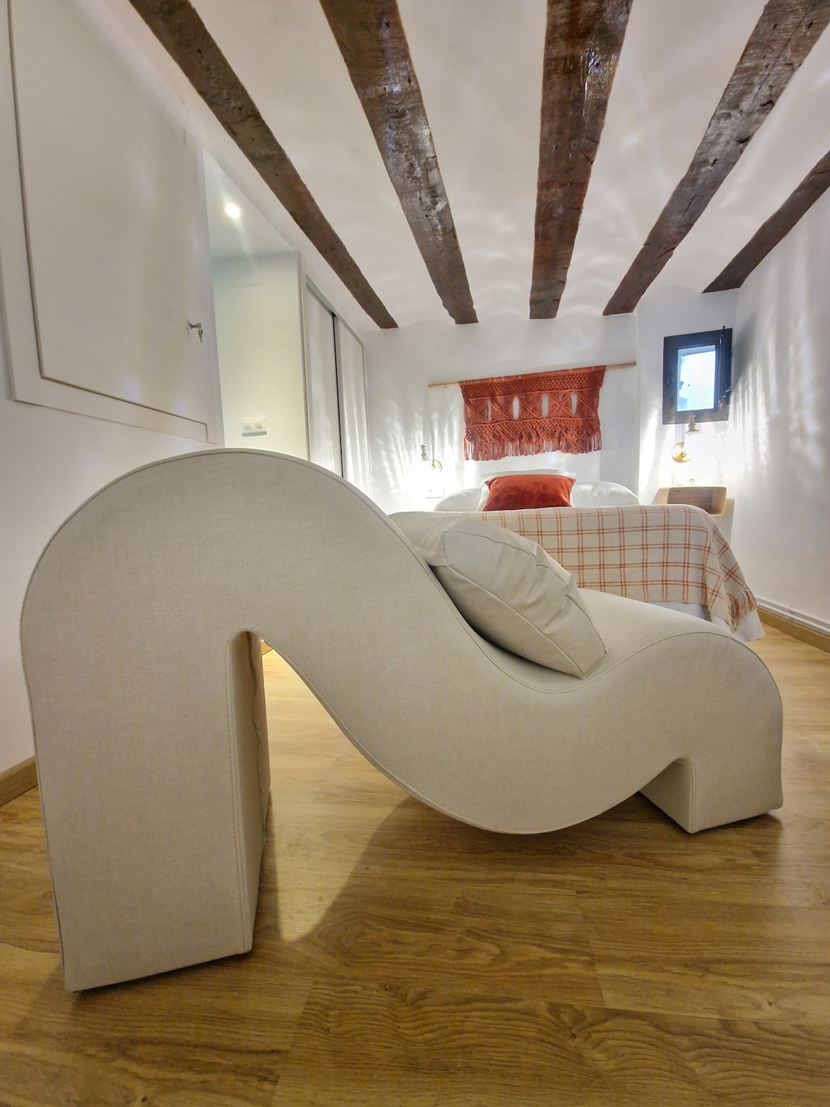 A uniquely designed seating piece with a sculptural wave shape is positioned in the foreground, featuring a soft fabric texture. In the background, a cozy bed with a checked coverlet is visible, along with exposed wooden beams adding character to the room.