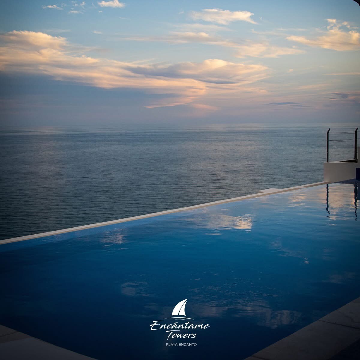 An infinity pool reflects the soft colors of the sunset over the ocean, creating a seamless view of the water. Gentle waves can be observed in the distance, and clouds are softly painted across the sky above.