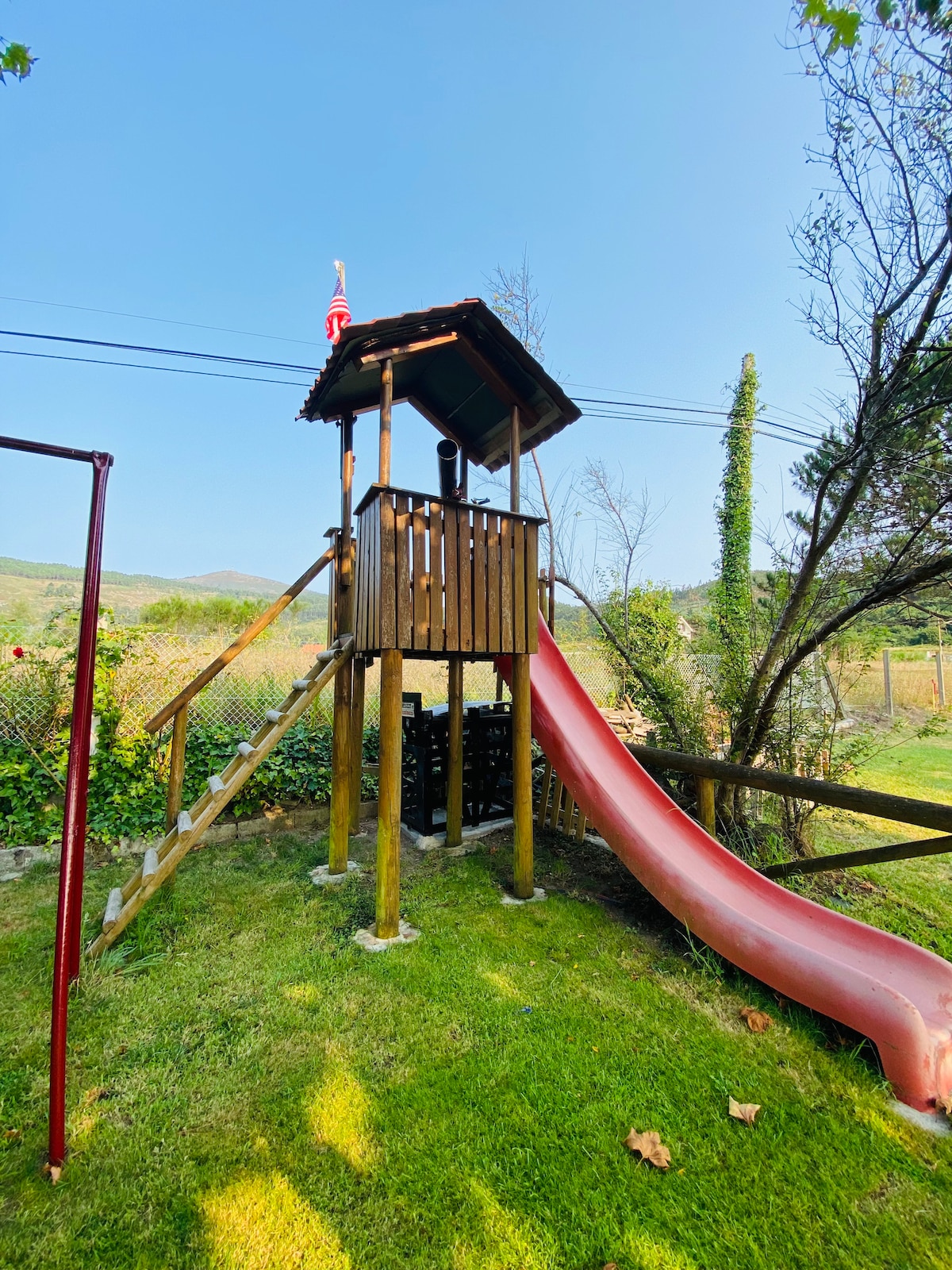 A wooden play structure is featured in the garden, complete with a slide and a small platform. Surrounding the play area, green grass and trees create a natural setting. The structure is designed for children, adding a playful element to the outdoor space.