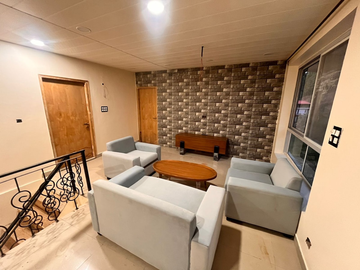 A welcoming living area is presented with light-colored seating arranged around a central wooden coffee table. A textured feature wall complements the comfortable ambiance, and large windows let in natural light, enhancing the room's spacious feel. Wooden doors are visible in the background.