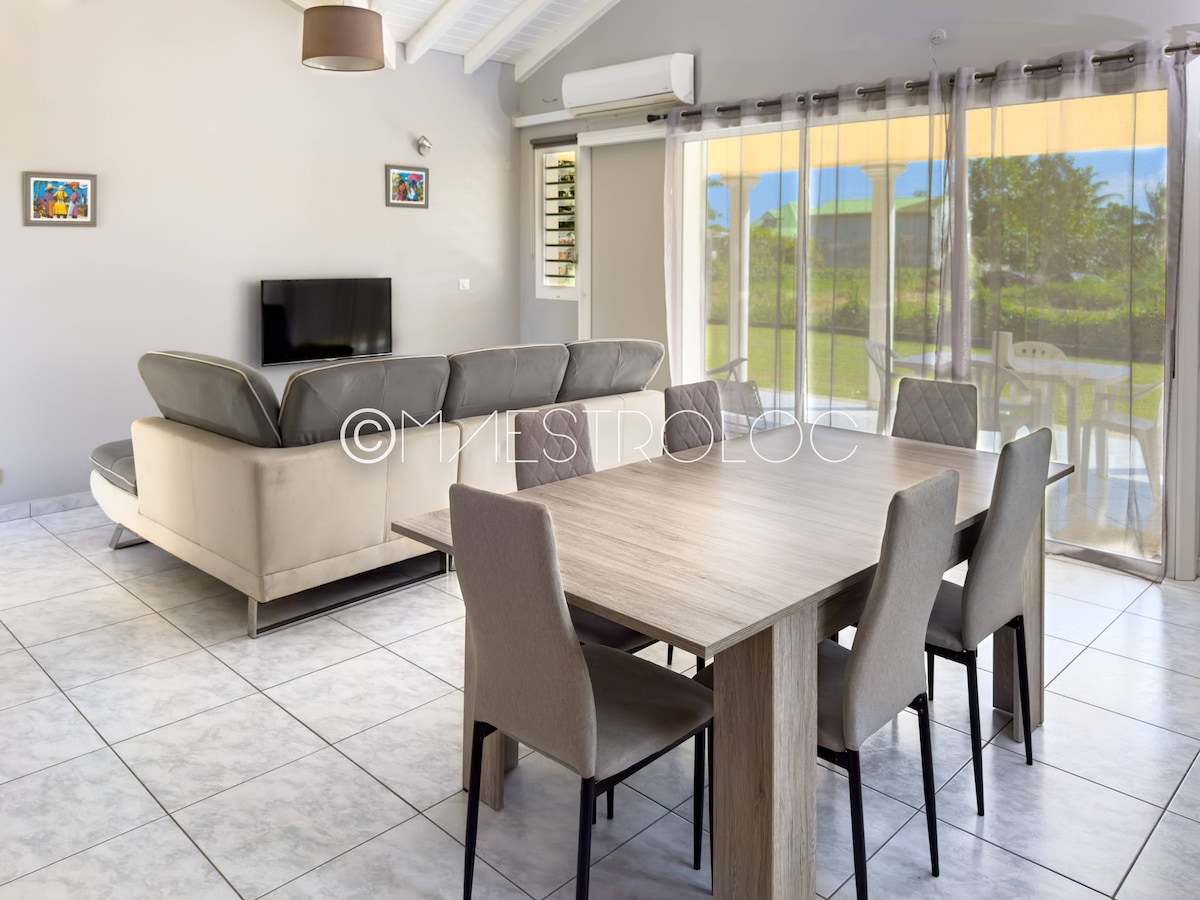 A light and airy living space is presented, featuring a comfortable grey sofa and a wooden dining table surrounded by six chairs. Natural light fills the room through large glass doors that lead to the terrace, offering a view of the garden.