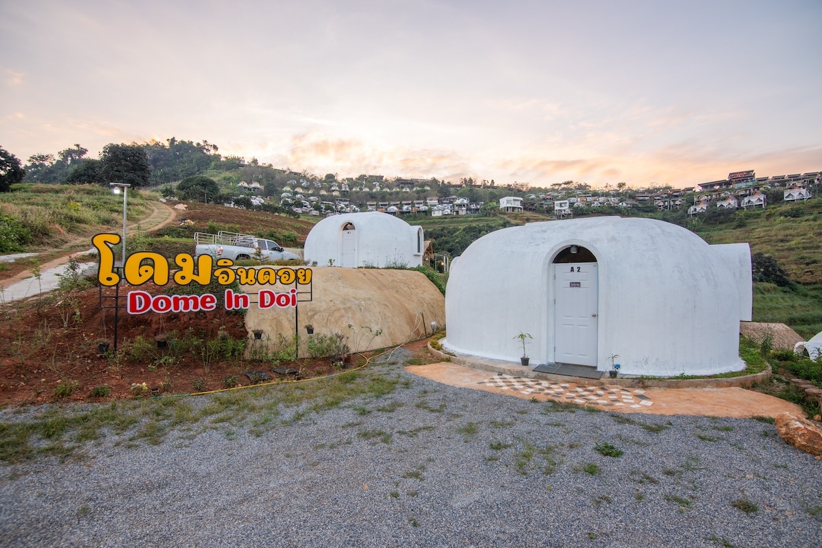 Dome-style accommodations are positioned on a hillside, surrounded by lush greenery. The exterior features smooth, white walls with a welcoming entrance. A gravel pathway leads to the domes, which are set against a backdrop of distant hills and scenic landscapes.