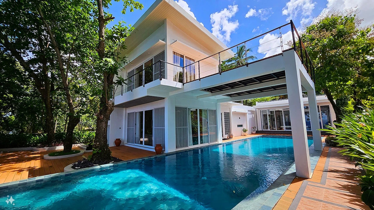 A modern two-story villa is surrounded by lush greenery, featuring large windows that provide ample natural light. An infinity pool runs along the side of the structure, reflecting the blue sky and white clouds above. Outdoor seating areas are visible on the smooth wooden deck.