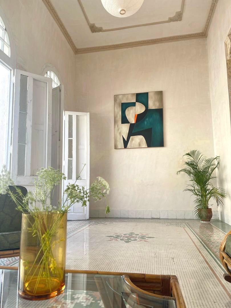 A bright and airy living area features a large abstract painting mounted on a light-colored wall. The space is enhanced by a vase of delicate white flowers and a lush green plant, providing a fresh and inviting look. Natural light streams through the open windows.