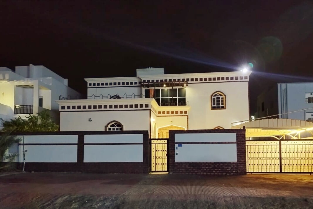 A modern guesthouse is showcased at night, highlighting its architectural features such as arched windows and a large balcony. The exterior is illuminated, revealing a welcoming entrance and a surrounding wall. The atmosphere conveys a sense of safety and tranquility.