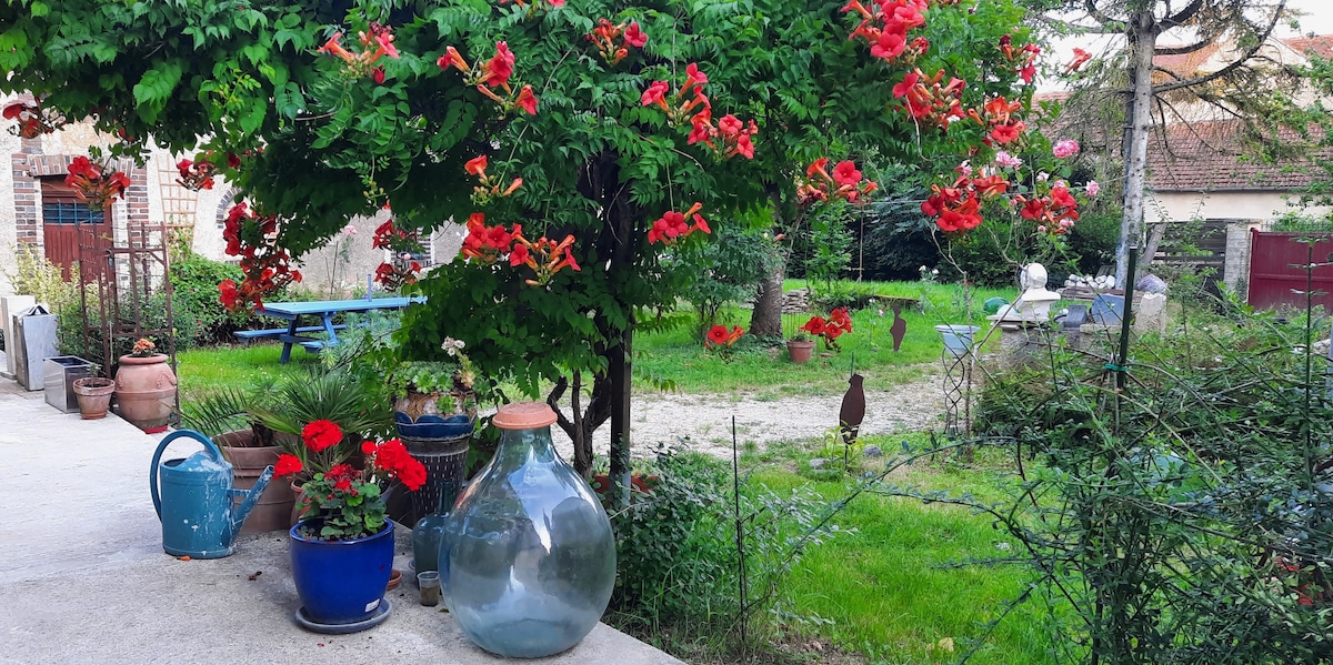 A vibrant garden setting showcases a variety of colorful flowers, including red blooms cascading from vines. A blue picnic table is visible in the background, along with decorative pots and a large glass jar placed on the ground, enhancing the natural landscape.