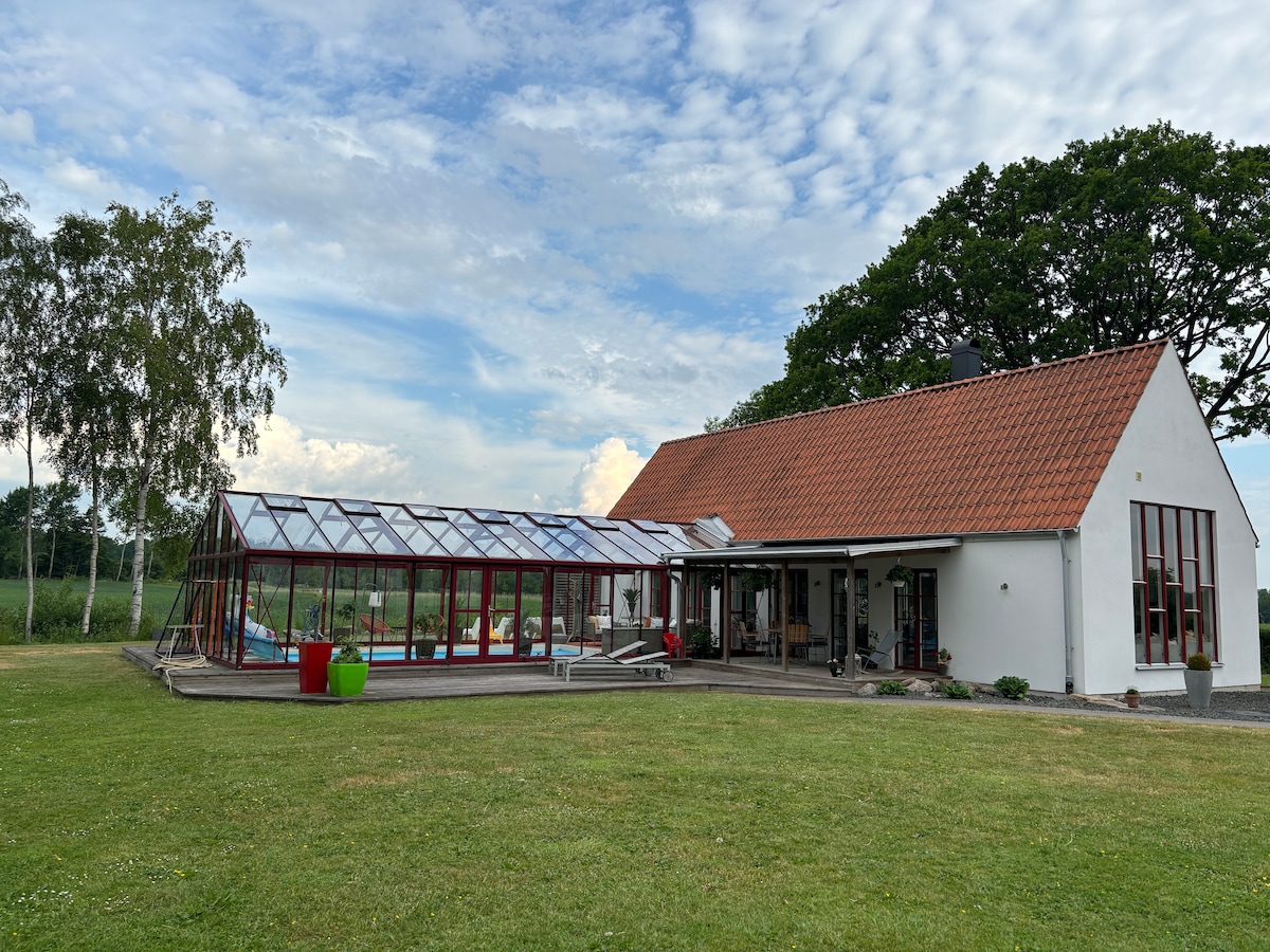 A modern structure displays a blend of white and red coloring, featuring a spacious greenhouse attached to one side. The property is surrounded by green grass and trees, with large windows illuminating the interior. A pool is visible within the greenhouse area.