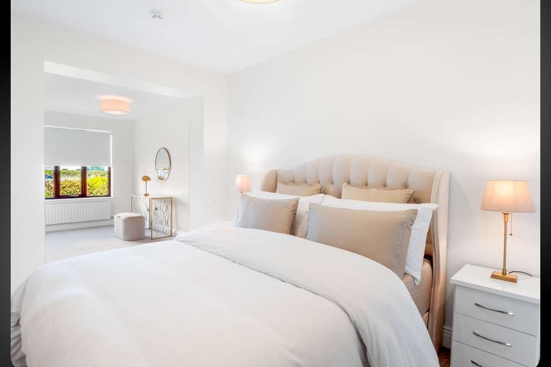 A serene bedroom is presented with a plush, beige tufted headboard and soft linens. Two bedside lamps provide gentle lighting, and a mirror reflects the natural light from the window. The space is airy, with light walls and a neutral color palette enhancing comfort.