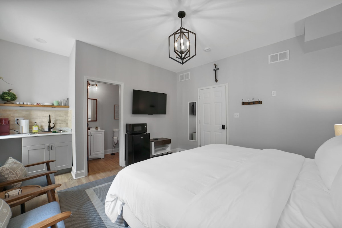 A modern bedroom features a spacious bed dressed in white linens. A wall-mounted television is visible across from a small kitchenette area. Soft lighting is provided by a decorative ceiling fixture. A bathroom entrance is seen in the background, framed by a pale wall.