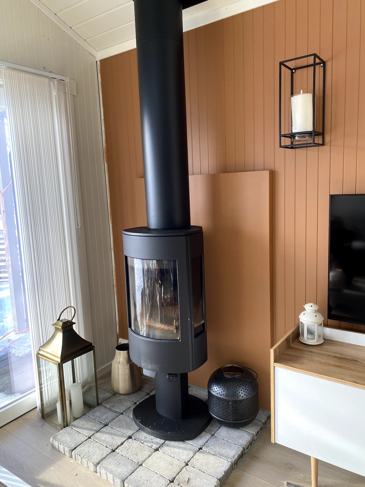 A modern wood stove stands against a warm, brown wall. Its glass front reveals flickering flames. To the side, a stone base supports the stove, while decorative lanterns and a sleek light fixture create a cozy ambiance. A minimalist media console is positioned nearby.