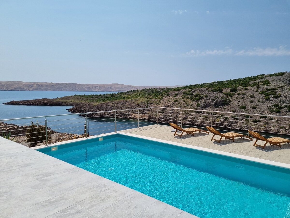 An outdoor area features a clear blue swimming pool surrounded by sun loungers on a tiled deck. The serene view showcases the coastline and rolling hills in the distance, reflecting the calmness of the water under a partially cloudy sky.