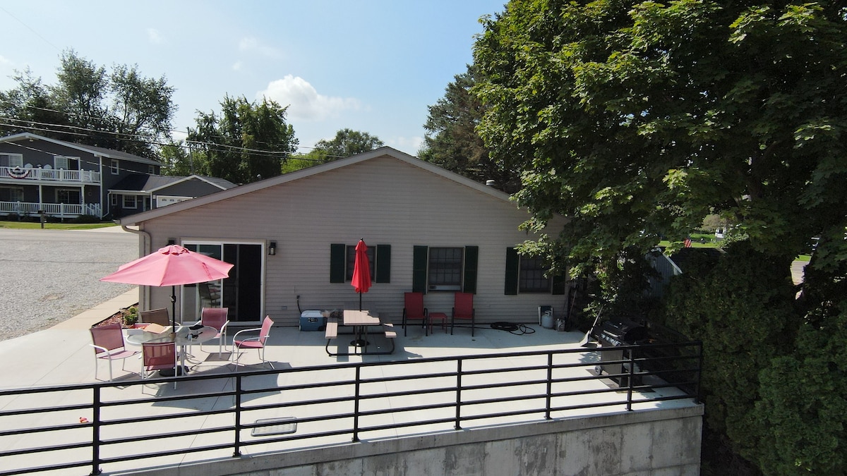 A spacious cement patio is highlighted, furnished with outdoor seating and umbrellas for shade. Greenery surrounds the area, enhancing the outdoor experience. The cottage exterior features a neutral color, with shuttered windows contributing to a balanced aesthetic.