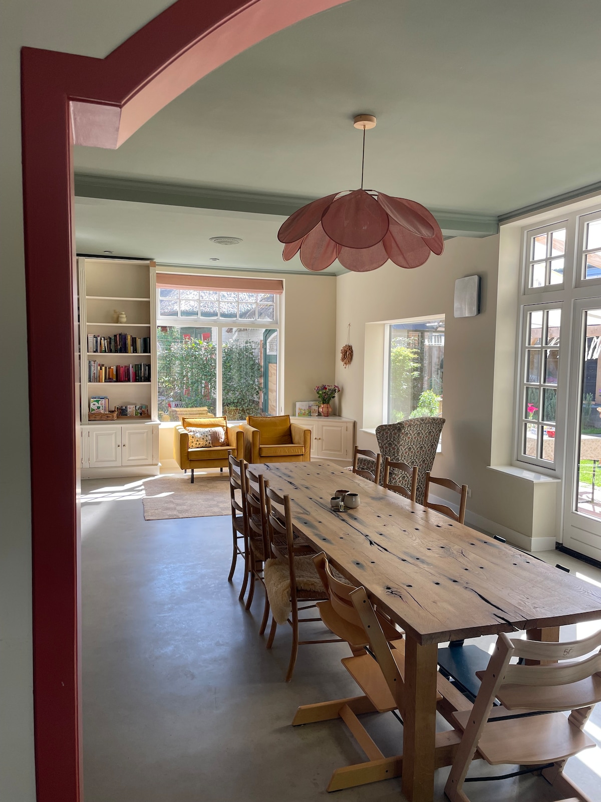 A spacious dining area features a long wooden table surrounded by multiple chairs. Large windows illuminate the space, showcasing views of the garden. A cozy reading corner with yellow chairs is visible, complemented by built-in shelves filled with books.