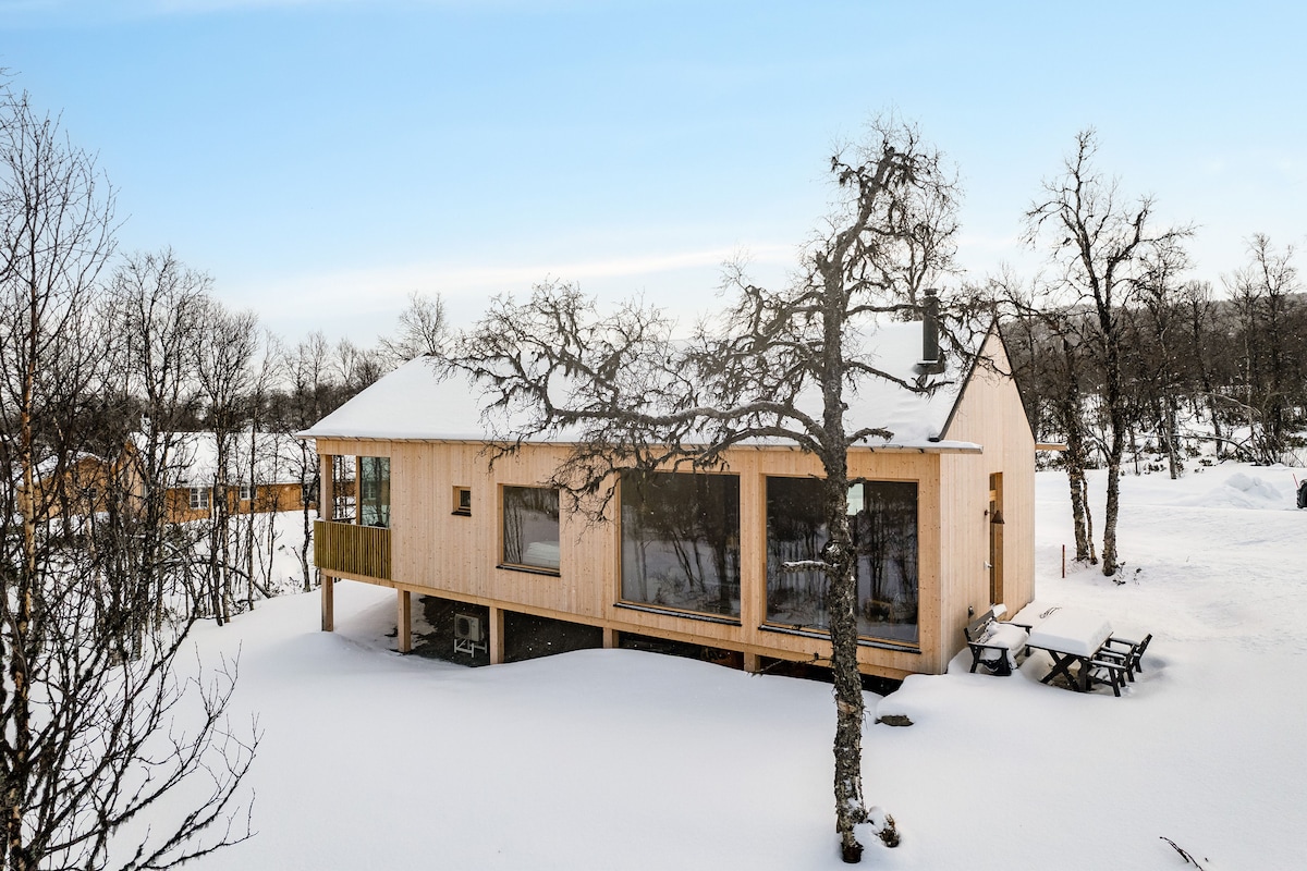 A modern cabin with a wooden exterior is surrounded by snow-covered terrain. Large windows allow for light to enter, providing views of the winter landscape. An outdoor seating area with a table is visible, complemented by nearby trees that enhance the natural setting.