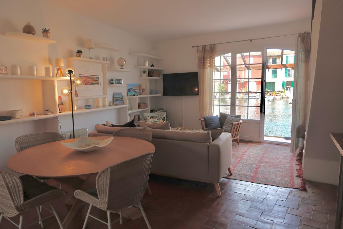 A well-arranged living area displays a round dining table with woven chairs, paired with a comfortable sofa. Light filters through large glass doors, providing access to an outdoor terrace. A television and decorative shelves complement the space, with views of the canal visible outside.