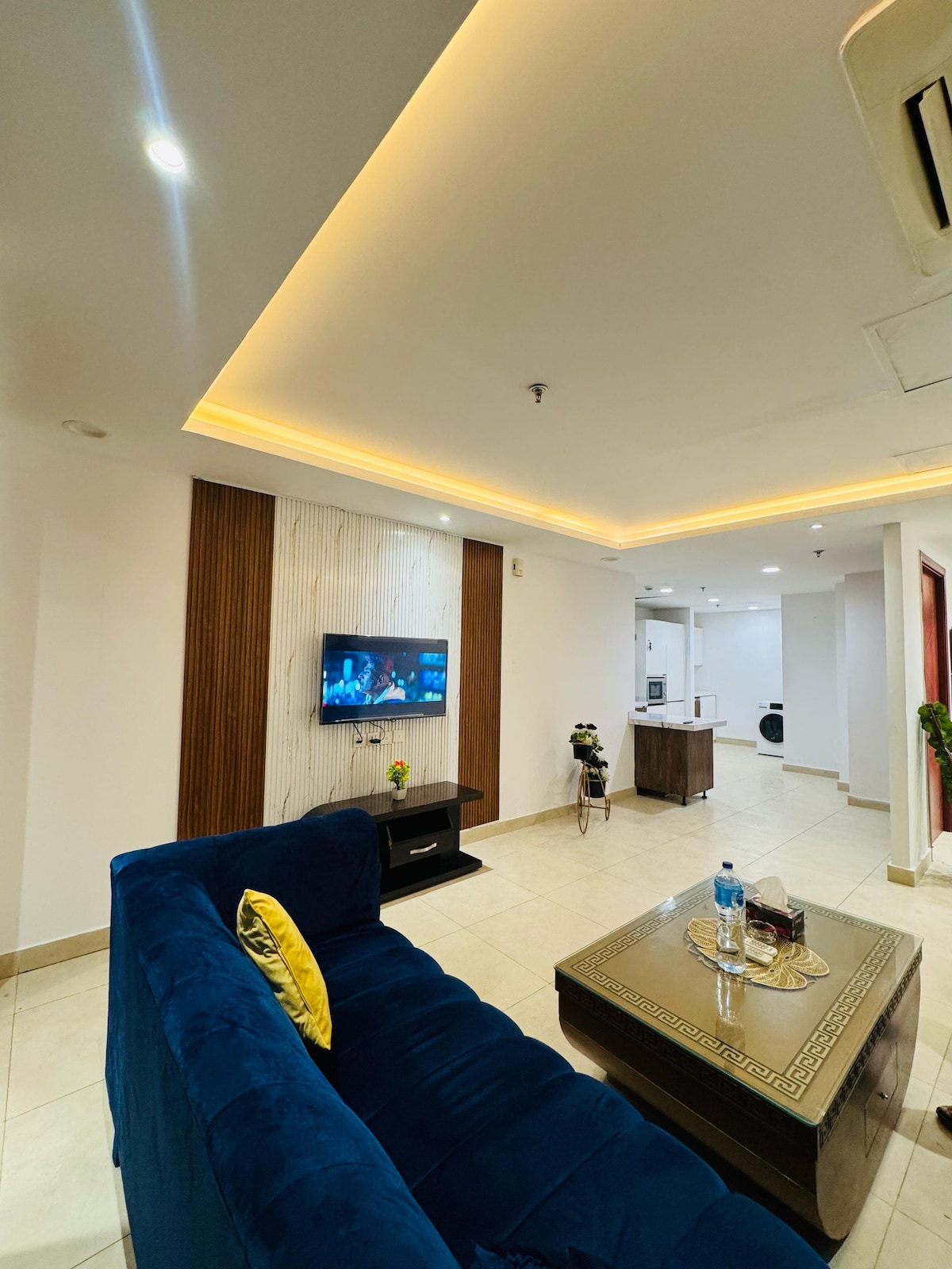 A modern living area features a plush blue sofa facing a wall-mounted smart TV. Soft lighting highlights a textured feature wall, while a glass coffee table showcases decorative items. A kitchenette and laundry area are visible in the background, providing convenience for guests.
