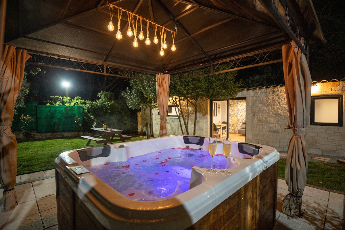 A private jacuzzi is nestled under a canopy, illuminated by soft hanging lights. The water is tinted with ambient colors and adorned with flower petals. A quaint garden setting surrounds the jacuzzi, creating a serene environment for relaxation.