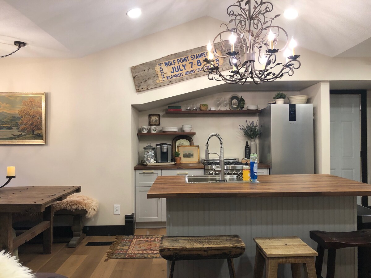 A warm kitchen area is highlighted by a wooden dining table and rustic stools. Above, an artistic chandelier enhances the space. A polished countertop complements modern appliances, while additional shelving provides storage for dishware and décor, contributing to the inviting atmosphere.